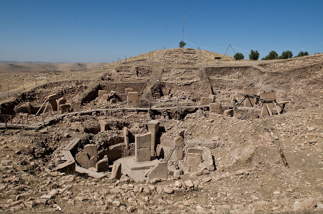 Visão panorâmica do sítio arqueológico de Göbekli Tepe na Turquia, mostrando grandes pilares megalíticos de pedra em formato de T dispostos em círculos sob uma estrutura moderna de proteção. Religião. Agricultura. Neolítico. Civilização. Arqueologia. Sociedade.