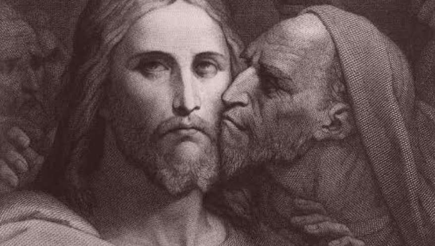 Gravura clássica em preto e branco retratando o Beijo de Judas em Jesus Cristo. Em primeiro plano, Jesus aparece de frente, com uma expressão serena, triste e olhar profundo. À sua direita, Judas, visto de perfil com feições duras, barba e a cabeça coberta por um manto, inclina-se para beijar o rosto de Cristo. Ao fundo, nas sombras, notam-se vagamente os rostos de outros homens.